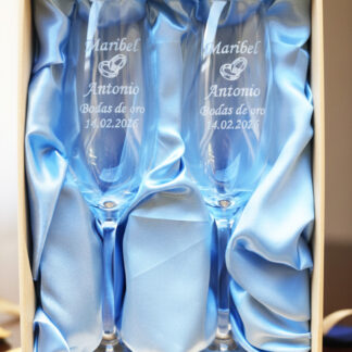 Dos copas de cava de cristal grabadas, presentadas en un estuche premium con forro de raso azul claro. Las copas llevan inscritos los nombres "Maribel y Antonio", un icono de alianzas, la frase "Bodas de oro" y la fecha "14.02.2026".