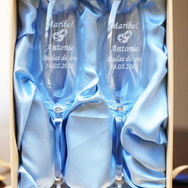 Dos copas de cava de cristal grabadas, presentadas en un estuche premium con forro de raso azul claro. Las copas llevan inscritos los nombres "Maribel y Antonio", un icono de alianzas, la frase "Bodas de oro" y la fecha "14.02.2026".