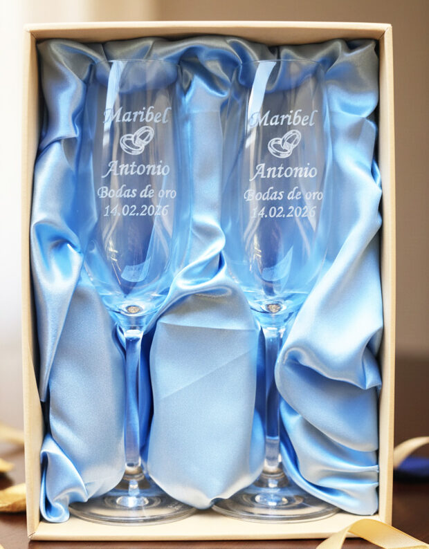Dos copas de cava de cristal grabadas, presentadas en un estuche premium con forro de raso azul claro. Las copas llevan inscritos los nombres "Maribel y Antonio", un icono de alianzas, la frase "Bodas de oro" y la fecha "14.02.2026".