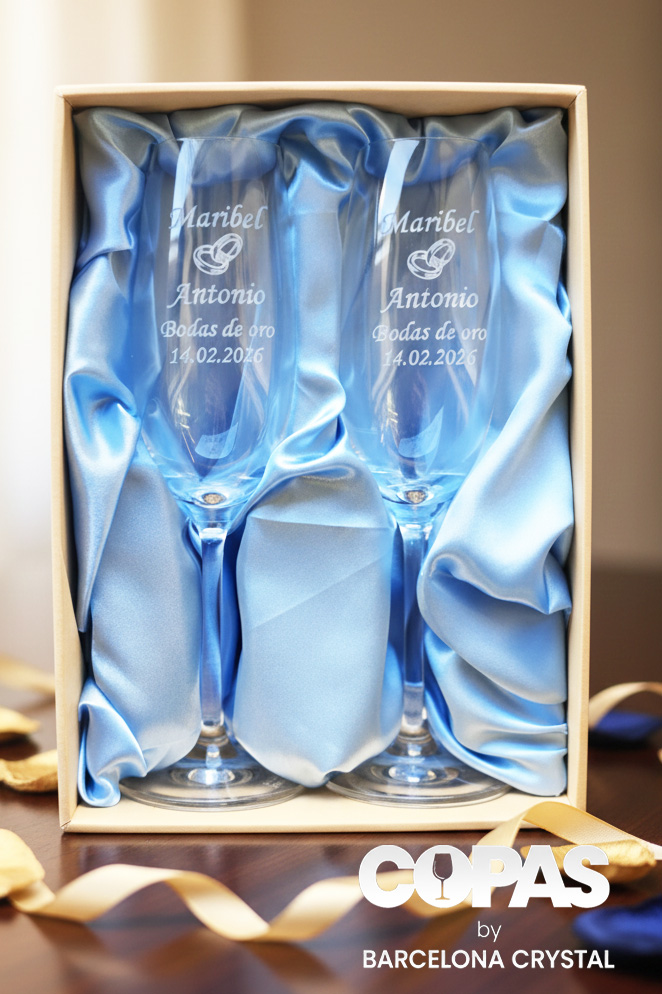 Dos copas de cava de cristal grabadas, presentadas en un estuche premium con forro de raso azul claro. Las copas llevan inscritos los nombres "Maribel y Antonio", un icono de alianzas, la frase "Bodas de oro" y la fecha "14.02.2026".