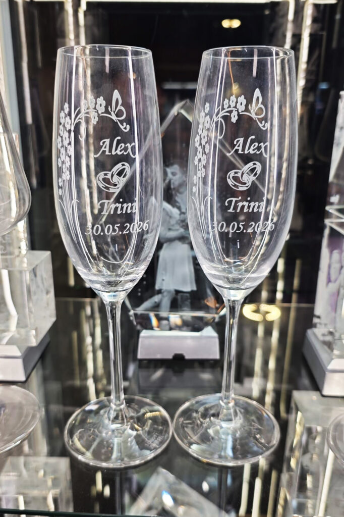 Pareja de copas de cava de cristal transparente con grabados personalizados, situadas sobre una superficie de cristal con un fondo gris neutro y limpio. Las copas muestran los nombres "Alex" y "Trini", un diseño de alianzas y la fecha "30.05.2026".