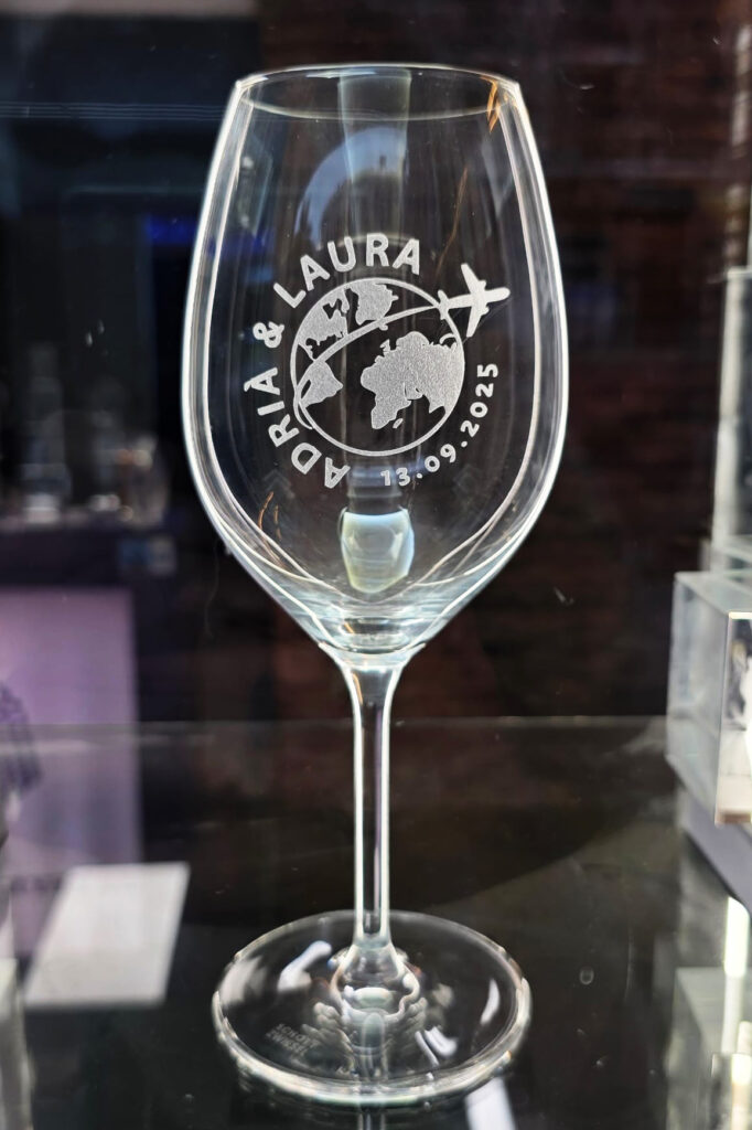 Copa de vino de cristal Zwiesel personalizada con un grabado del mapa del mundo, un avión siguiendo una ruta punteada, los nombres "Adrià & Laura" y la fecha "13.09.2025". La copa está sobre una superficie oscura reflectante con un fondo negro elegante.