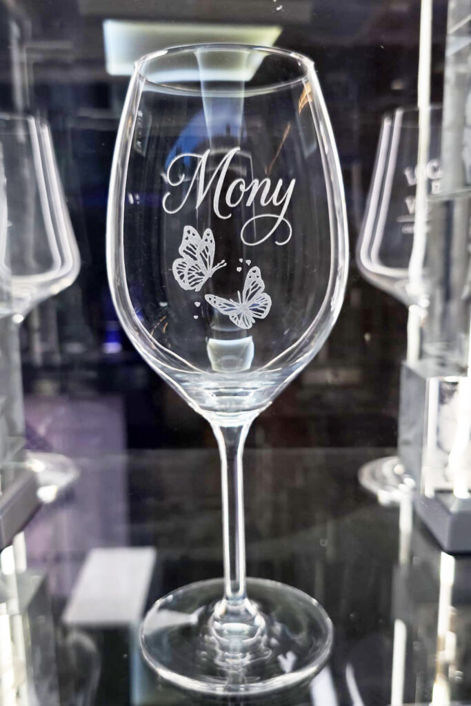 Una copa de vino de cristal Zwiesel personalizada con el nombre "Mony" grabado en caligrafía elegante y dos mariposas detalladas debajo del nombre. La copa está en primer plano con un fondo de cristalería desenfocado.