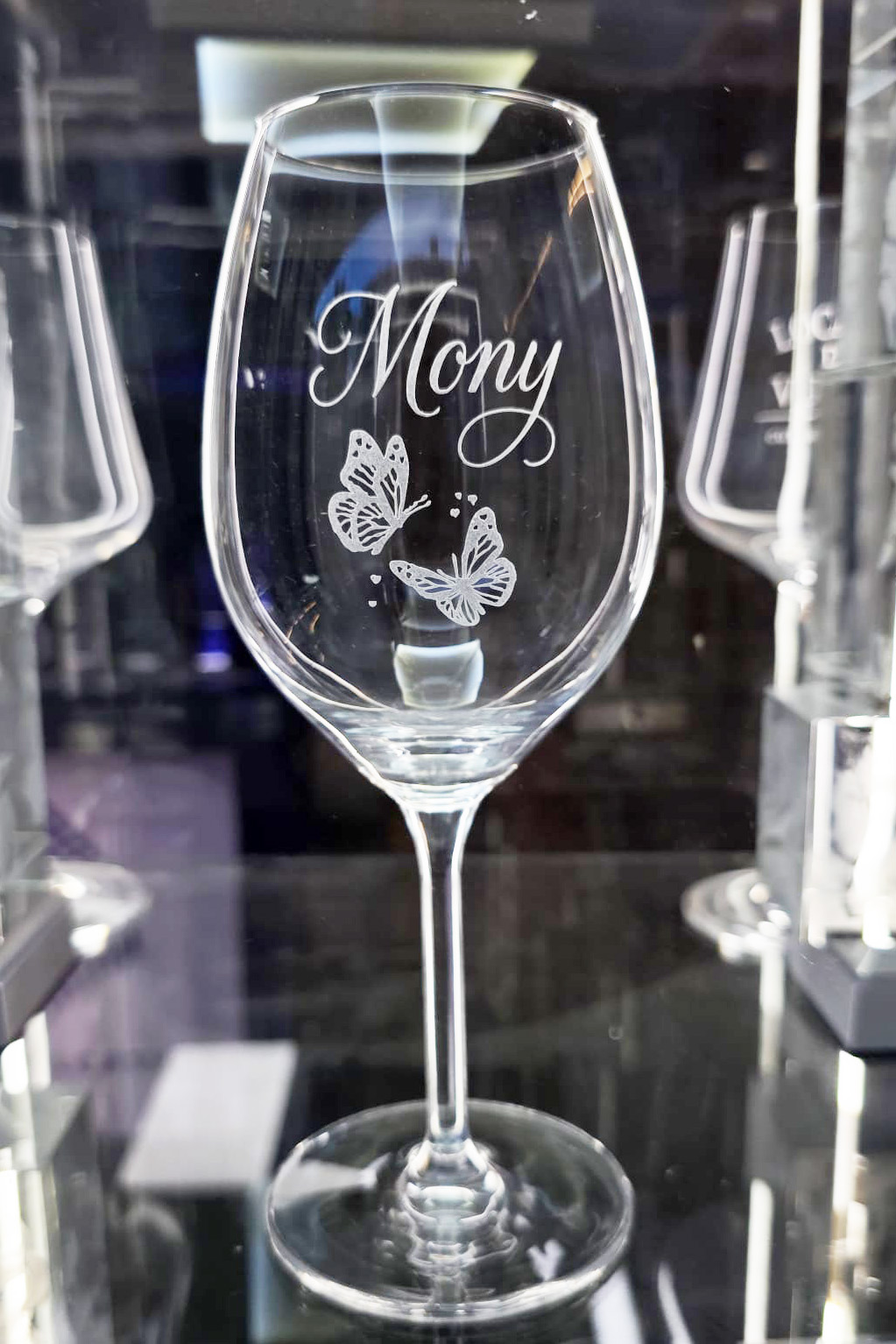 Una copa de vino de cristal Zwiesel personalizada con el nombre "Mony" grabado en caligrafía elegante y dos mariposas detalladas debajo del nombre. La copa está en primer plano con un fondo de cristalería desenfocado.