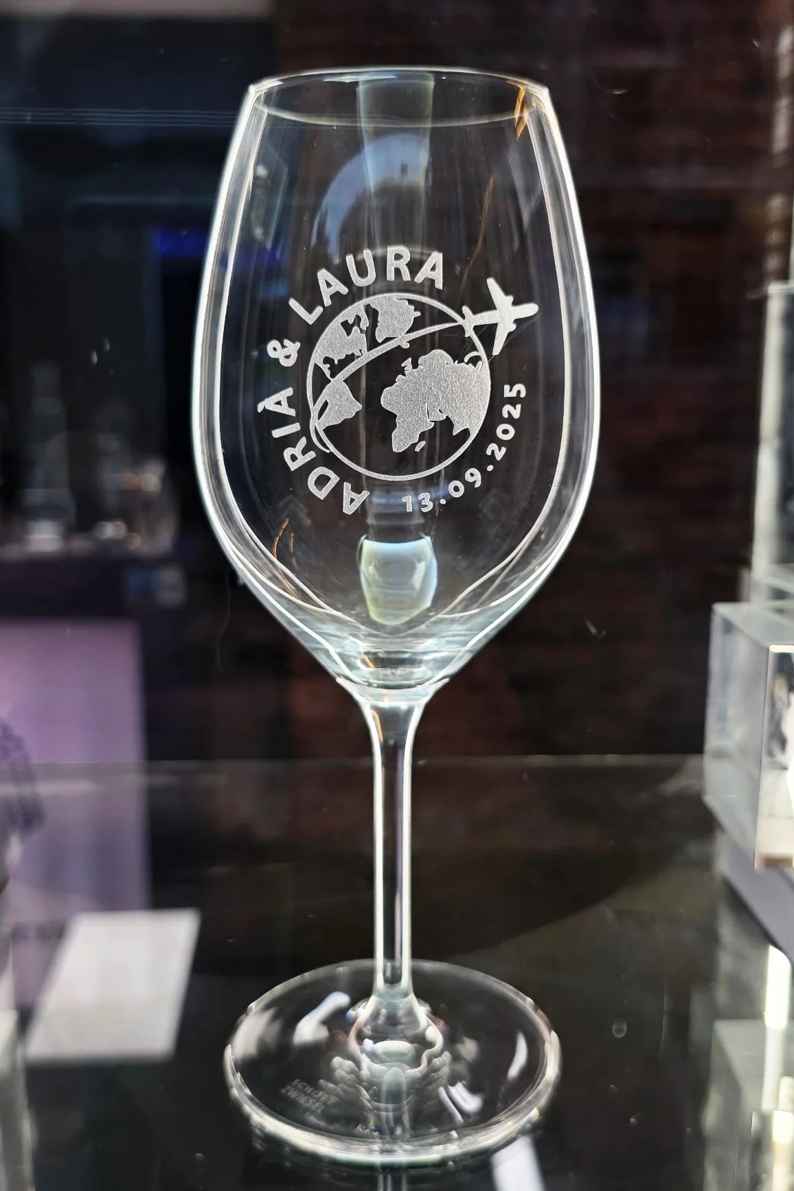 Copa de vino de cristal Zwiesel personalizada con un grabado del mapa del mundo, un avión siguiendo una ruta punteada, los nombres "Adrià & Laura" y la fecha "13.09.2025". La copa está sobre una superficie oscura reflectante con un fondo negro elegante.